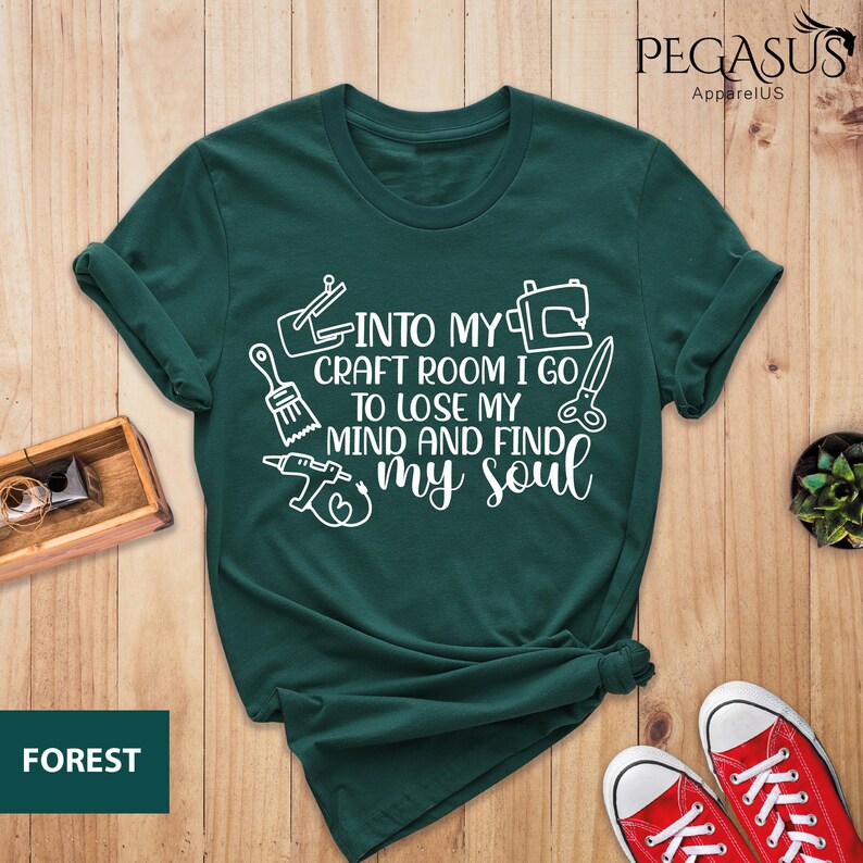 K&ouml;nnte beinhalten: Ein dunkelgr&uuml;nes T-Shirt mit wei&szlig;em Text, der "Into my craft room I go to lose my mind and find my soul" lautet. Der Text ist von Bildern von Bastelwerkzeugen umgeben, darunter ein Pinsel, eine N&auml;hmaschine und eine S&auml;ge.