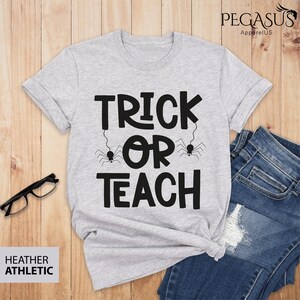 May include: A heather gray t-shirt with the words "TRICK OR TEACH" in black, with spider graphics. The shirt is paired with blue jeans and glasses on a wooden surface. The shirt is labeled "HEATHER ATHLETIC".