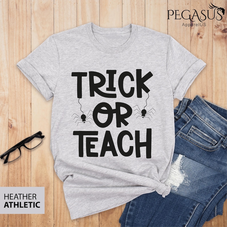 May include: A heather gray t-shirt with the words "TRICK OR TEACH" in black, with spider graphics. The shirt is paired with blue jeans and glasses on a wooden surface. The shirt is labeled "HEATHER ATHLETIC".