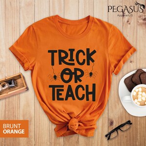 May include: Burnt orange t-shirt with the text "TRICK OR TEACH" in black, with spider graphics. The shirt is knotted at the bottom. The brand name "PEGASUS ApparelUS" is visible in the top right corner.