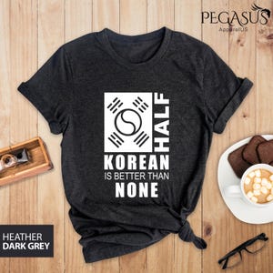 May include: A dark heather grey t-shirt with a white graphic design. The graphic features the Korean flag and the text "Half Korean is better than none".