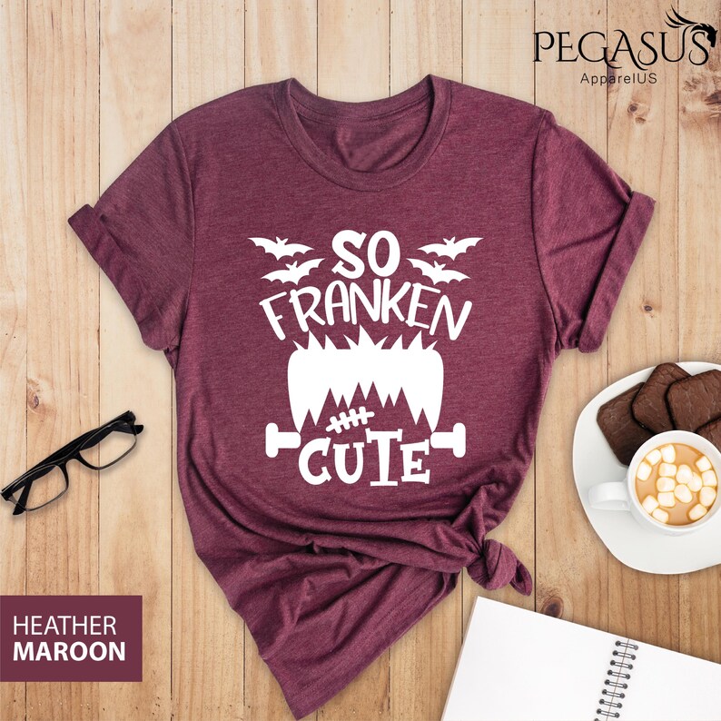 Pu&ograve; includere: T-shirt color bordeaux m&eacute;lange con la scritta bianca "SO FRANKEN CUTE", un disegno di mostro di Frankenstein e pipistrelli. La maglietta &egrave; esposta su una superficie di legno con un paio di occhiali, un taccuino e una tazza di caff&egrave;.