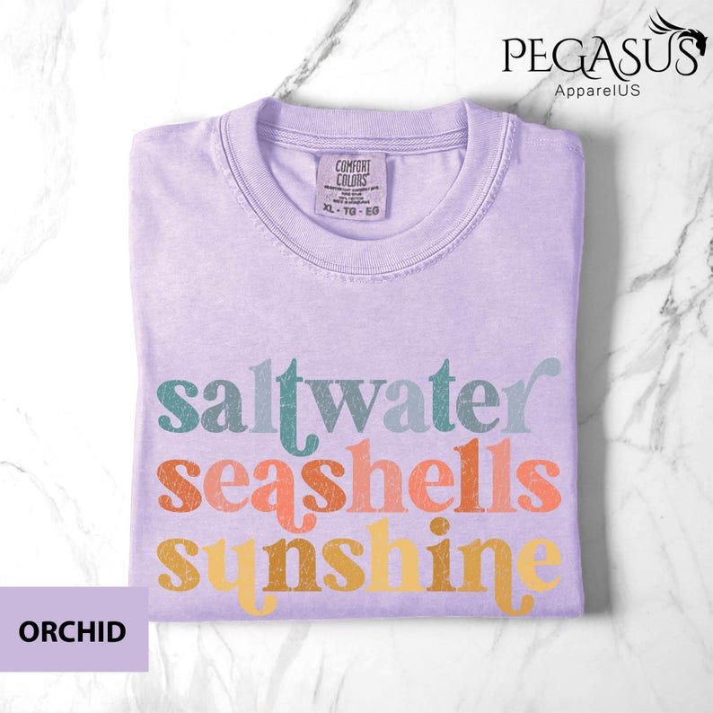 May include: Orchid-colored t-shirt with the words "saltwater seashells sunshine" in teal, coral, and yellow lettering. The shirt has a crew neck and a Comfort Colors tag. The shirt is folded neatly on a marble surface.