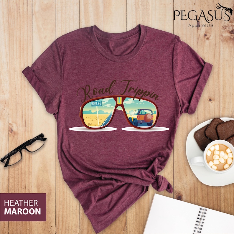 Peut inclure: T-shirt en marron chin&eacute; avec un motif de lunettes de soleil refl&eacute;tant une sc&egrave;ne de road trip. Le texte "Road Trippin'" est au-dessus des lunettes de soleil.