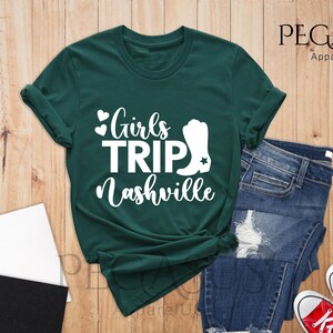 May include: Forest green t-shirt with the text "Girls Trip Nashville" in white. The design includes hearts and a cowboy boot graphic. The shirt is paired with blue jeans and red sneakers, suggesting a casual, travel-themed outfit.