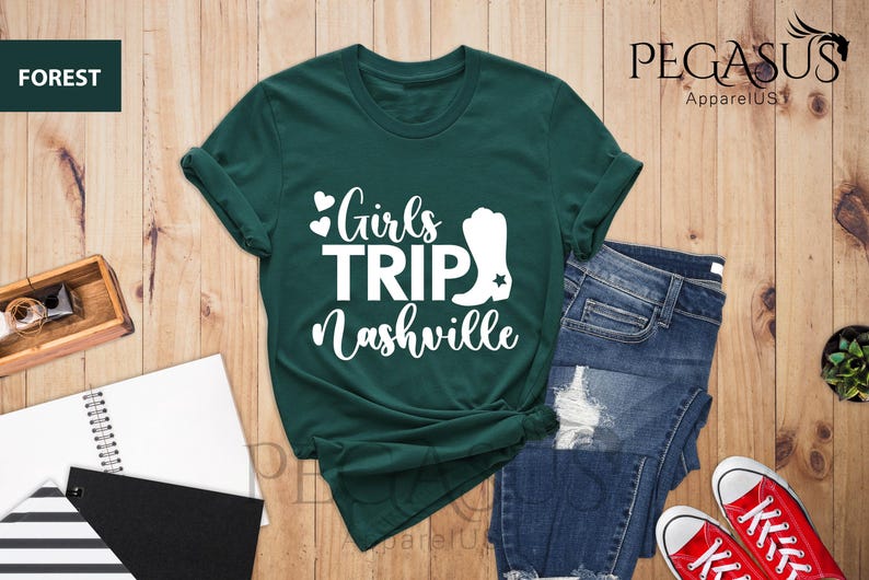 May include: Forest green t-shirt with the text "Girls Trip Nashville" in white. The design includes hearts and a cowboy boot graphic. The shirt is paired with blue jeans and red sneakers, suggesting a casual, travel-themed outfit.