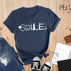 May include: A heather navy blue t-shirt with the word "SMILE" spelled out using dental tools. The shirt is laid flat on a wooden surface with a pair of jeans, a notebook, and a pair of glasses.