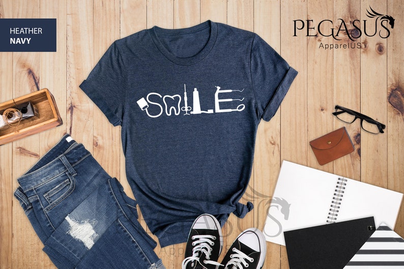 May include: A heather navy blue t-shirt with the word "SMILE" spelled out using dental tools. The shirt is laid flat on a wooden surface with a pair of jeans, a notebook, and a pair of glasses.