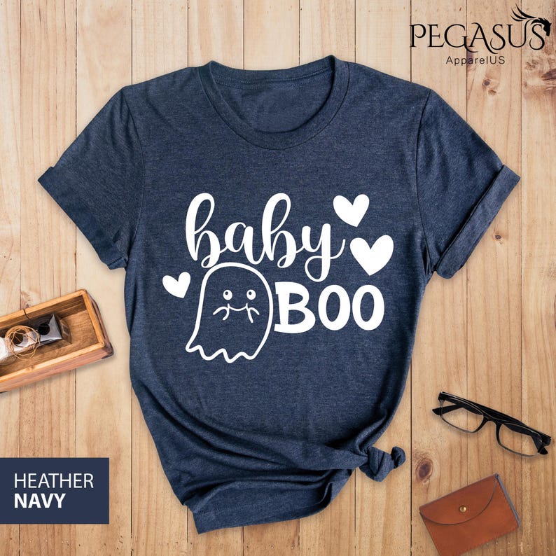May include: Heather navy blue t-shirt with the words "baby BOO" in white script and a ghost graphic. The shirt has a crew neck and short sleeves. The text "HEATHER NAVY" is in a navy blue rectangle in the bottom left corner.