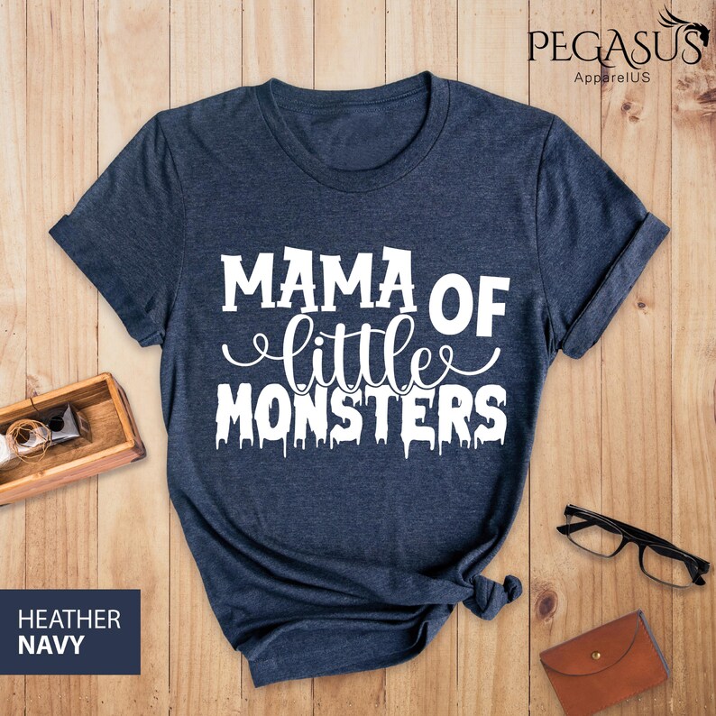 May include: Heather navy blue t-shirt with the text "MAMA OF little MONSTERS" in white. The shirt is displayed on a wooden surface with a small wooden box, glasses, and a wallet.