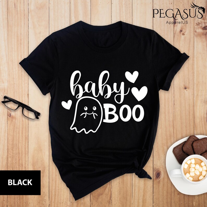 May include: Black t-shirt with the text "baby BOO" in white, featuring a ghost graphic and heart accents. The shirt is from Pegasus ApparelUS. A pair of black glasses and a cup of coffee with cookies are on the wooden surface.