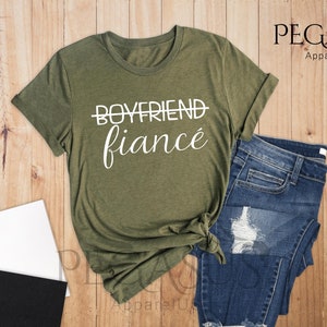 May include: A green t-shirt with the text "Boyfriend" crossed out and "fianc&eacute;" written below it. The shirt is on a wooden surface with a pair of jeans, a notebook, and a cup of coffee.
