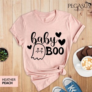May include: Heather peach t-shirt with the words "baby BOO" in black script and a ghost graphic. The shirt has a crew neck and short sleeves. The shirt is on a wooden surface with a pair of blue sneakers and a plate of cookies.