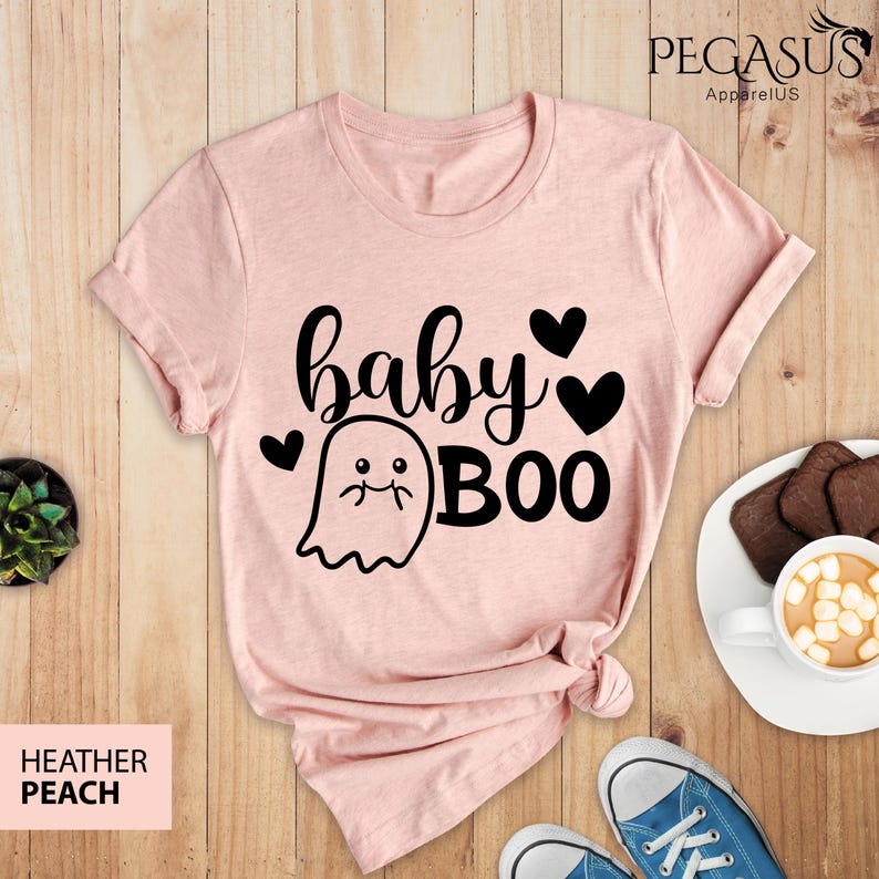 May include: Heather peach t-shirt with the words "baby BOO" in black script and a ghost graphic. The shirt has a crew neck and short sleeves. The shirt is on a wooden surface with a pair of blue sneakers and a plate of cookies.