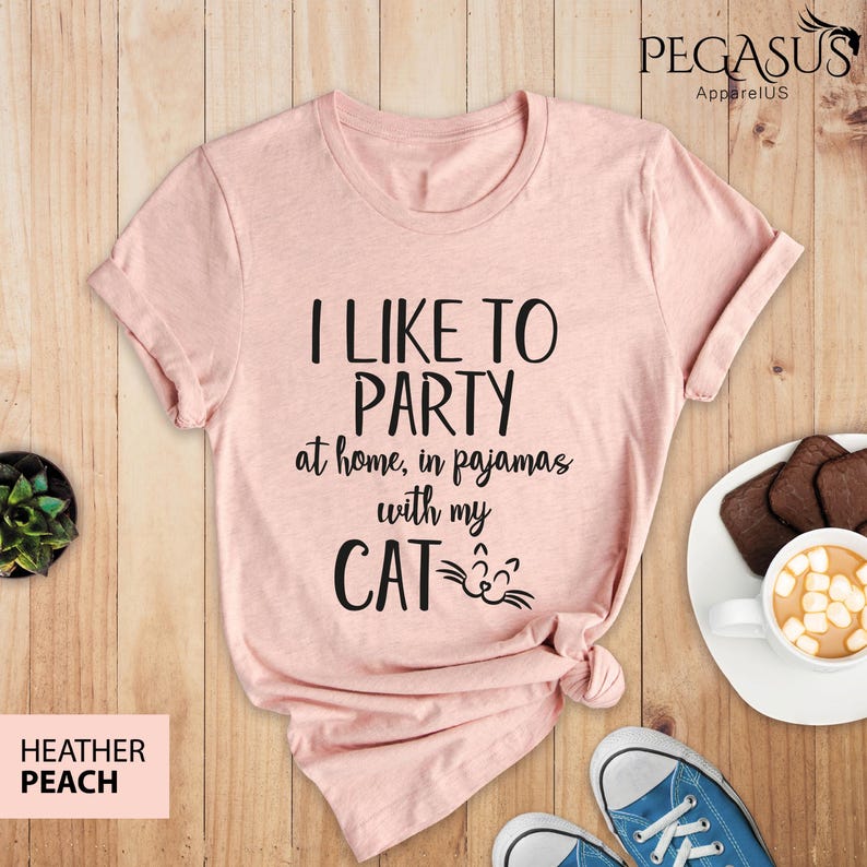 Op de afbeelding: Een roze T-shirt met de tekst "Ik vind het leuk om te feesten thuis, in pyjama met mijn kat" in zwarte letters. De kat heeft een schattig gezicht met snorharen.