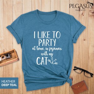Op de afbeelding: Een tealblauw t-shirt met de tekst "Ik vind het leuk om te feesten thuis, in pyjama met mijn kat" en een kattengezicht-afbeelding.