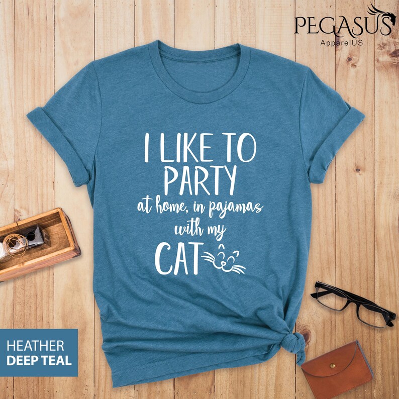 Op de afbeelding: Een tealblauw t-shirt met de tekst "Ik vind het leuk om te feesten thuis, in pyjama met mijn kat" en een kattengezicht-afbeelding.
