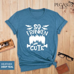 Pu&ograve; includere: Una maglietta verde acqua scuro con la scritta bianca "SO FRANKEN CUTE", con un design di mostro di Frankenstein e pipistrelli. La maglietta &egrave; ripiegata sulle maniche e annodata in basso. Le parole "Heather Deep Teal" sono in un riquadro blu.