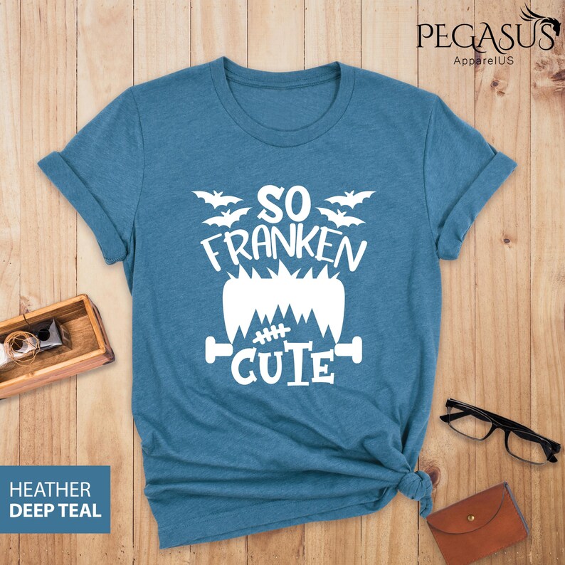 Pu&ograve; includere: Una maglietta verde acqua scuro con la scritta bianca "SO FRANKEN CUTE", con un design di mostro di Frankenstein e pipistrelli. La maglietta &egrave; ripiegata sulle maniche e annodata in basso. Le parole "Heather Deep Teal" sono in un riquadro blu.