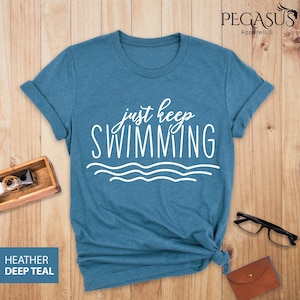 May include: A teal blue t-shirt with the text "just keep swimming" in white lettering. The text is in a cursive font and is accompanied by a wavy line design.