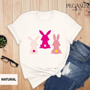 May include: White t-shirt featuring three Easter bunny silhouettes in pink and patterned pink. The middle bunny is solid pink, while the others have patterned designs. The word "NATURAL" is printed on a white rectangle.