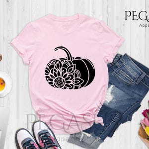 May include: A pink t-shirt featuring a black graphic of a pumpkin with a mandala design. The shirt is laid out on a white wooden surface with a pair of jeans, sunglasses, a cup of tea, a laptop, and a bouquet of flowers.