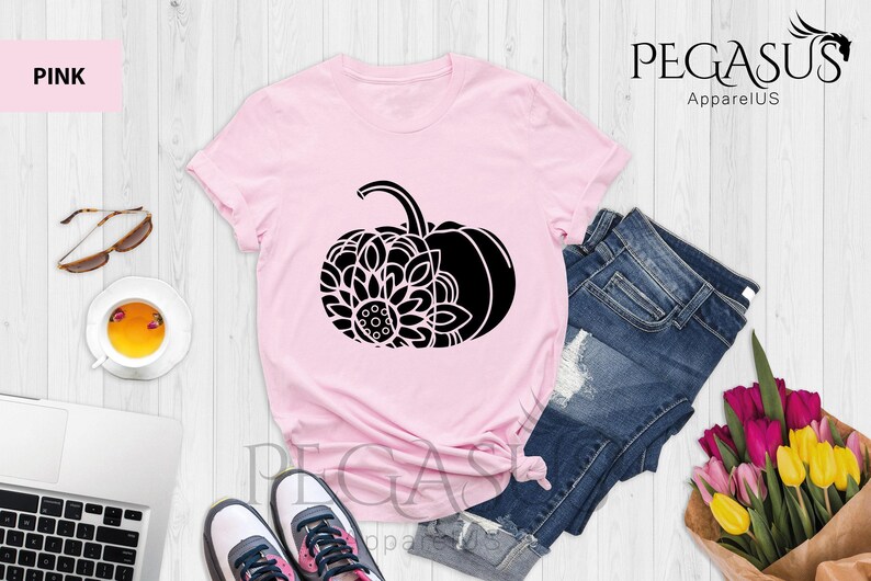 May include: A pink t-shirt featuring a black graphic of a pumpkin with a mandala design. The shirt is laid out on a white wooden surface with a pair of jeans, sunglasses, a cup of tea, a laptop, and a bouquet of flowers.