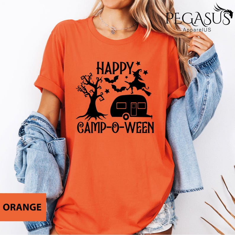 May include: Orange t-shirt with a black graphic that reads "HAPPY CAMP-O-WEEN". The graphic features a tree, bats, a witch on a broom, stars, and a camper. The shirt is a short-sleeved crew neck.