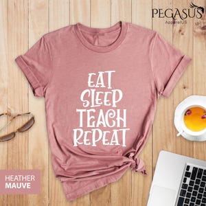 May include: A heather mauve t-shirt with the text "Eat Sleep Teach Repeat" printed in white.
