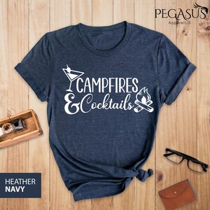 May include: Heather navy blue t-shirt with the words "Campfires & Cocktails" in white script. The design includes a martini glass and a campfire graphic. The shirt is displayed on a wooden surface.