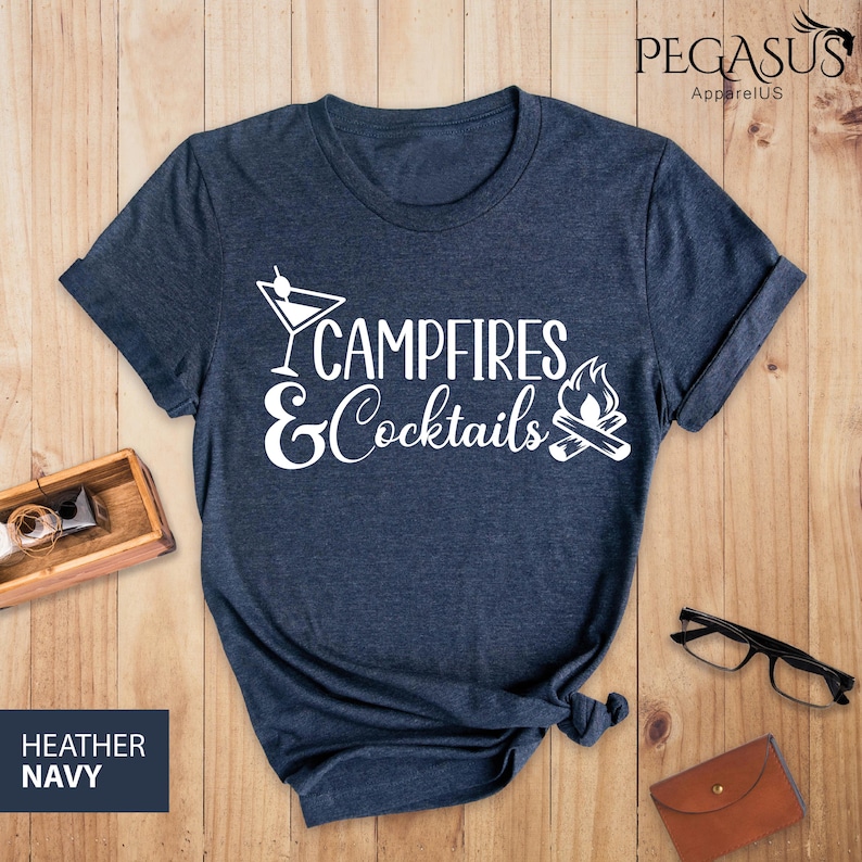 May include: Heather navy blue t-shirt with the words "Campfires & Cocktails" in white script. The design includes a martini glass and a campfire graphic. The shirt is displayed on a wooden surface.