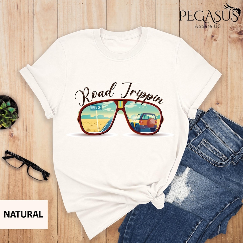 Peut inclure: T-shirt blanc avec un graphique de lunettes de soleil refl&eacute;tant une route d&eacute;sertique avec une voiture roulant sur la Route 66. Le texte "Road Trippin'" est au-dessus des lunettes de soleil.