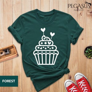 May include: A dark green t-shirt with a white cupcake graphic. The cupcake has white frosting, hearts, and stars. The text "PEGASUS ApparelUS" is in the top right corner of the image. The text "FOREST" is in the bottom left corner of the image.