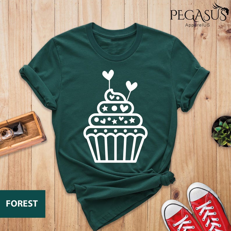 May include: A dark green t-shirt with a white cupcake graphic. The cupcake has white frosting, hearts, and stars. The text "PEGASUS ApparelUS" is in the top right corner of the image. The text "FOREST" is in the bottom left corner of the image.