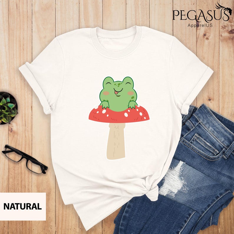 May include: White t-shirt with a green frog sitting on a red and white mushroom. The frog is smiling and has its tongue sticking out.