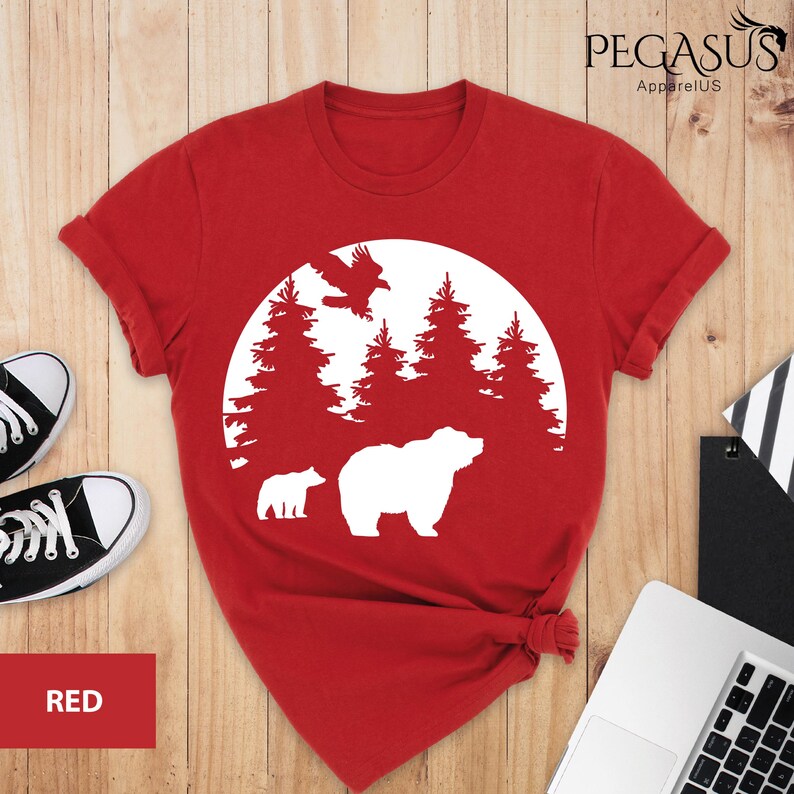 Puede incluir: Camiseta roja con un gr&aacute;fico blanco de un oso, un cachorro, un p&aacute;jaro y &aacute;rboles en un c&iacute;rculo. El texto "PEGASUS ApparelUS" est&aacute; en la esquina superior derecha.