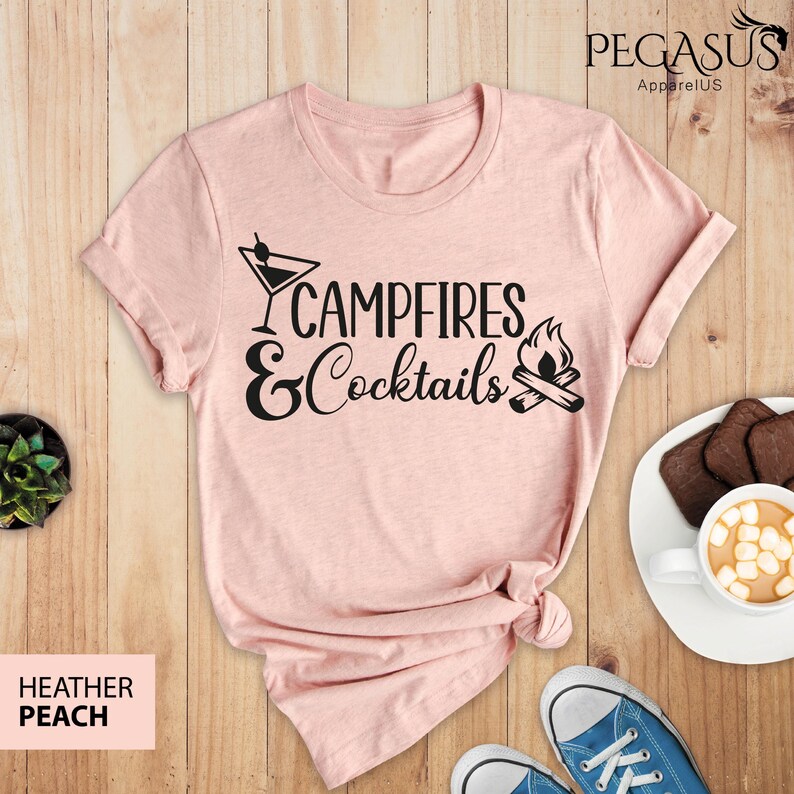 May include: A heather peach t-shirt with the words "Campfires & Cocktails" in black script and a martini glass and campfire graphic. The shirt is styled with blue sneakers and a plate of cookies.