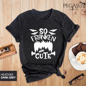Pu&ograve; includere: T-shirt grigio scuro melange con grafica bianca che dice "So Franken Cute" con pipistrelli e un design di mostro di Frankenstein. La maglietta &egrave; piegata su una superficie di legno con una tazza di cioccolata calda e occhiali.