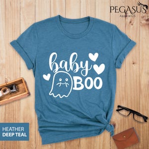 May include: Teal-colored t-shirt with the words "baby BOO" in white script and a ghost graphic. The shirt has rolled-up sleeves and a knotted hem. The text "HEATHER DEEP TEAL" is in a blue box in the lower left corner.