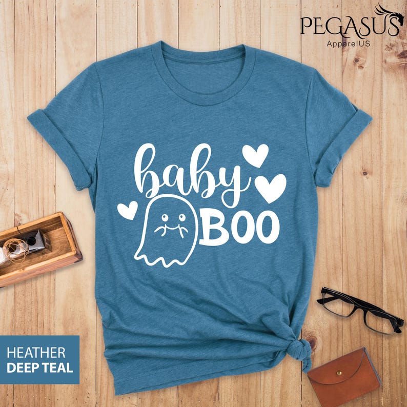 May include: Teal-colored t-shirt with the words "baby BOO" in white script and a ghost graphic. The shirt has rolled-up sleeves and a knotted hem. The text "HEATHER DEEP TEAL" is in a blue box in the lower left corner.