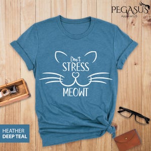 Op de afbeelding: Een tealblauw t-shirt met een witte kattenkop-afbeelding en de tekst "Don't Stress Meowt".