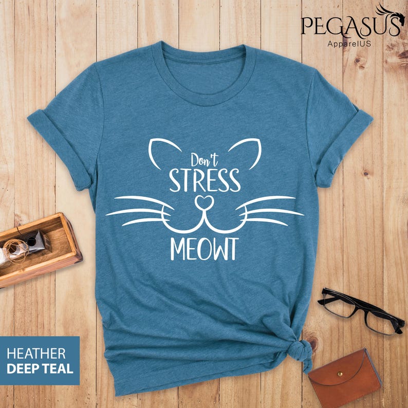 Op de afbeelding: Een tealblauw t-shirt met een witte kattenkop-afbeelding en de tekst "Don't Stress Meowt".