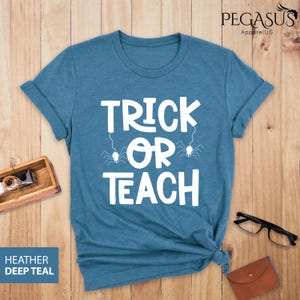 May include: Teal-colored t-shirt with the words "TRICK OR TEACH" in white, with spider graphics. The shirt is folded at the bottom. The words "HEATHER DEEP TEAL" are in a blue box in the lower left corner. The brand name "PEGASUS ApparelUS" is in the upper right corner.