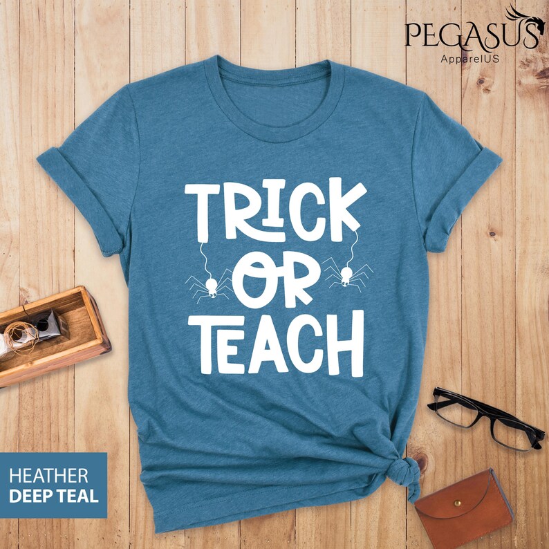 May include: Teal-colored t-shirt with the words "TRICK OR TEACH" in white, with spider graphics. The shirt is folded at the bottom. The words "HEATHER DEEP TEAL" are in a blue box in the lower left corner. The brand name "PEGASUS ApparelUS" is in the upper right corner.