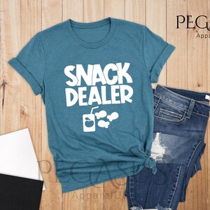 Peut inclure: Un t-shirt bleu sarcelle avec le texte "Snack Dealer" en blanc. Le t-shirt a un graphique d'une bo&icirc;te de jus avec une pomme et une paille, et trois bulles de poisson.