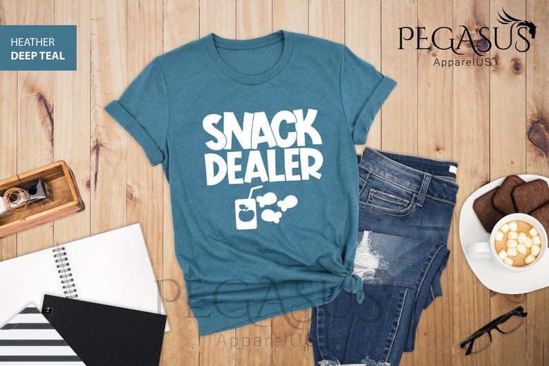 Peut inclure: Un t-shirt bleu sarcelle avec le texte "Snack Dealer" en blanc. Le t-shirt a un graphique d'une bo&icirc;te de jus avec une pomme et une paille, et trois bulles de poisson.