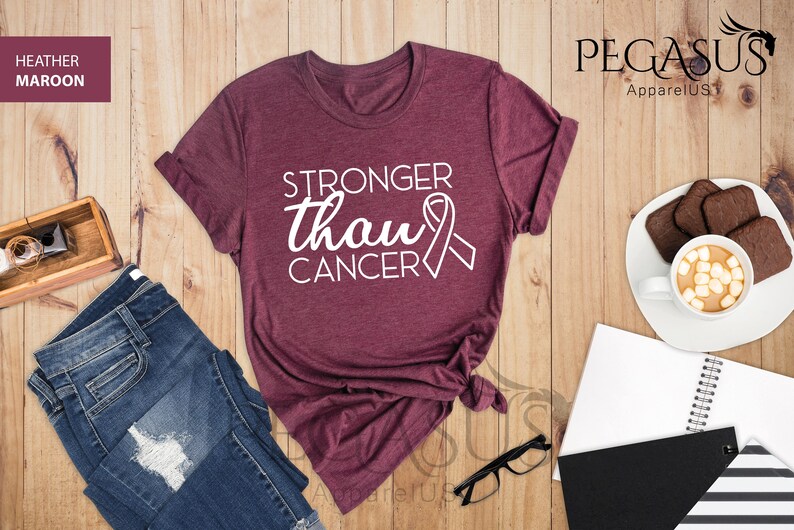 May include: A heather maroon t-shirt with the text "Stronger than Cancer" and a white ribbon graphic. The shirt is folded and displayed on a wooden surface with other items.