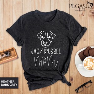 May include: Dark heather grey t-shirt with a white outline of a Jack Russell Terrier's face and the words "JACK RUSSEL MOM" in white script. The shirt is folded and knotted at the bottom. The text "HEATHER DARK GREY" is in a box in the lower left corner.