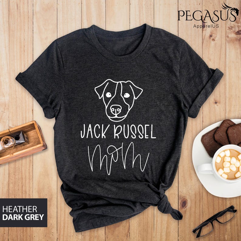 May include: Dark heather grey t-shirt with a white outline of a Jack Russell Terrier's face and the words "JACK RUSSEL MOM" in white script. The shirt is folded and knotted at the bottom. The text "HEATHER DARK GREY" is in a box in the lower left corner.