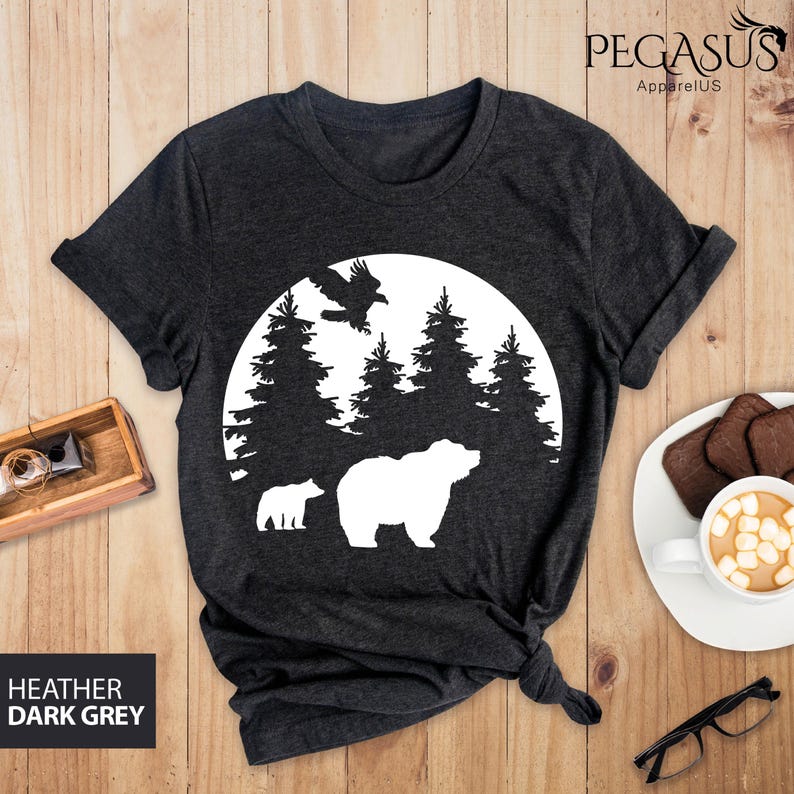 Puede incluir: Una camiseta gris oscuro jaspeada con un dise&ntilde;o de silueta blanca de un oso, un p&aacute;jaro y &aacute;rboles en un c&iacute;rculo. El dise&ntilde;o recuerda a una escena de bosque.
